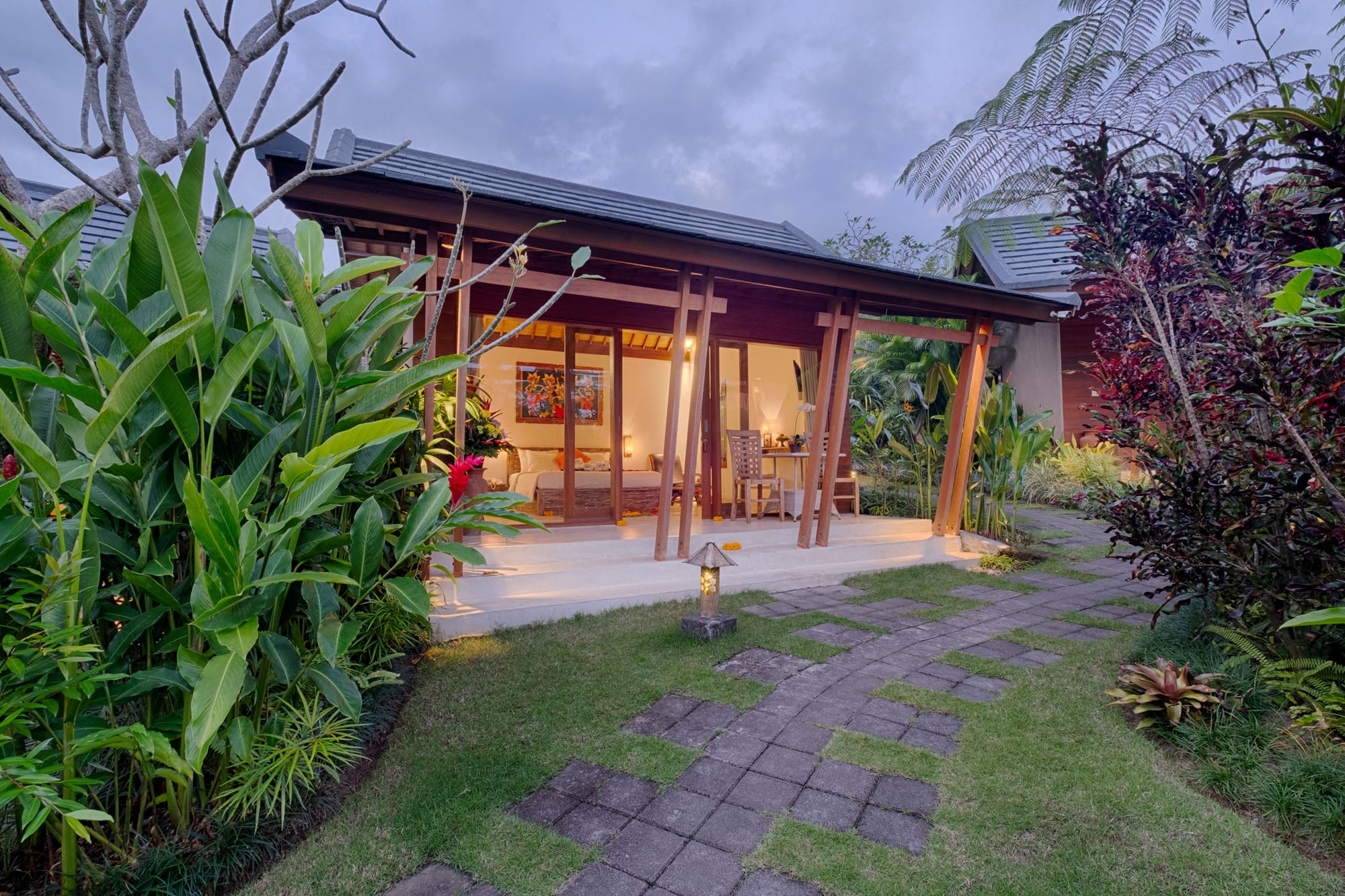 Cozy traditional Balinese wooden villa structure surrounded by tropical plants under a twilight sky at TegalTis Villas Payangan.